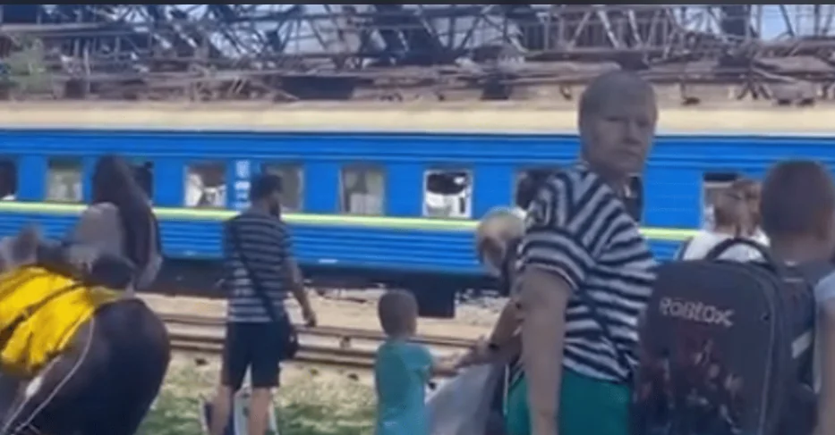 Люди з дітьми біля синього поїзда з розбитими вікнами та пошкодженою контактною мережею